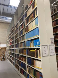 A tall metal bookshelf, with 8 bays, each with 10 shelves full of books, towers towards an industrial roof.