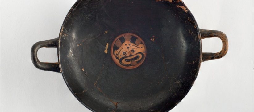 Image shows a round black-figure cup that has two handles. In the centre of the cup, an image of Medusa's face is visible and she has her tongue out. The rest of the cup is black, with the exception of a slight wear.